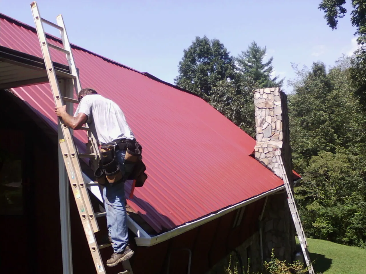 Roofer on a ladder beside a red metal roof and stone chimney — Soffit & Fascia Repair by Roofing Brothers in Pattison